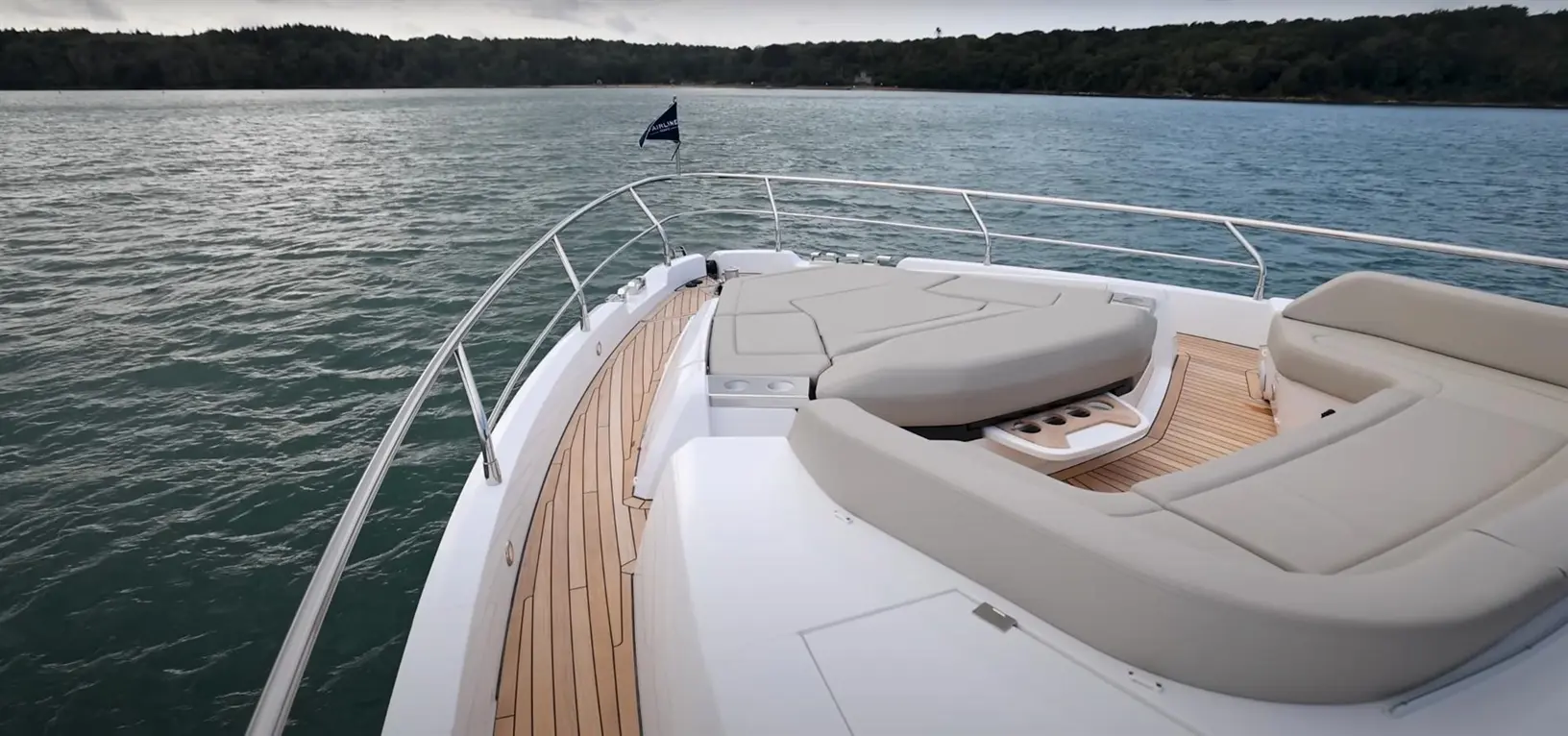 A luxury boat's bow features comfortable seating and teak flooring, with calm blue water and lush greenery in the background.